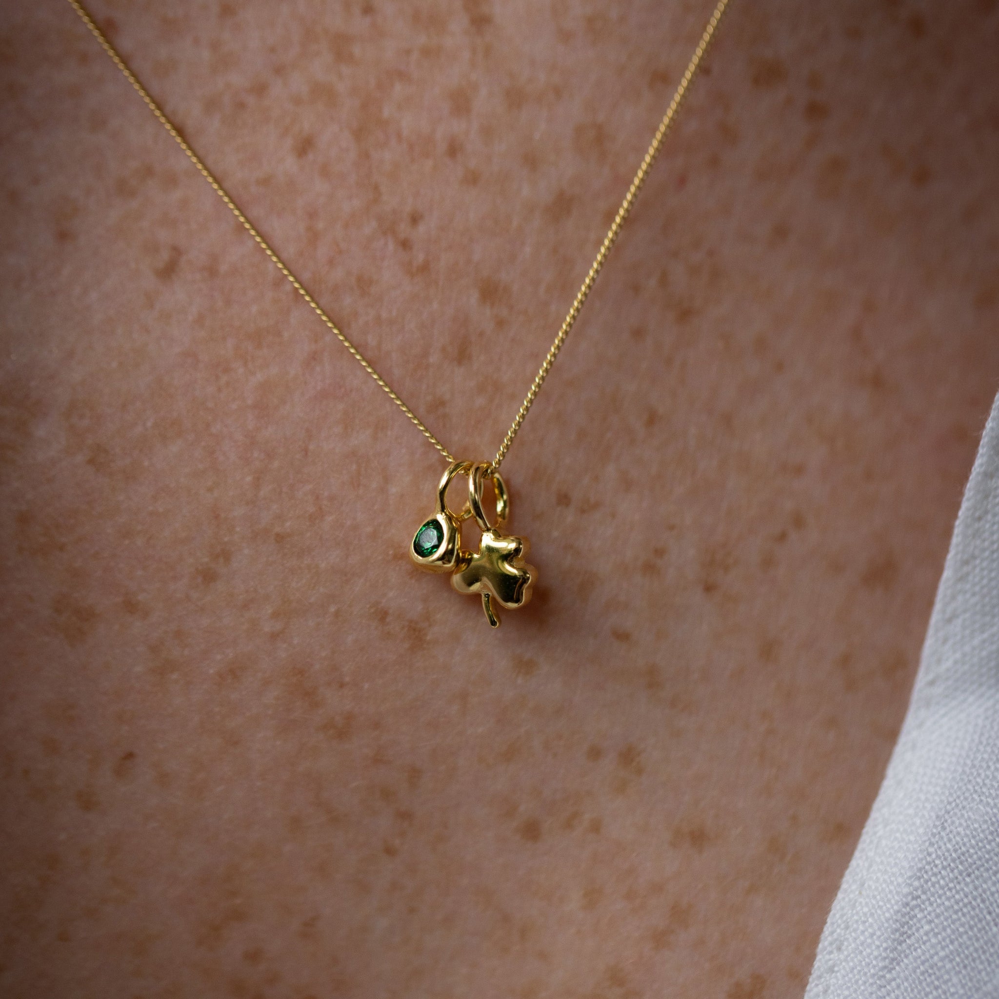 A close-up of a delicate gold chain necklace worn on a person's chest, featuring a small gold charm with green gemstone accents.