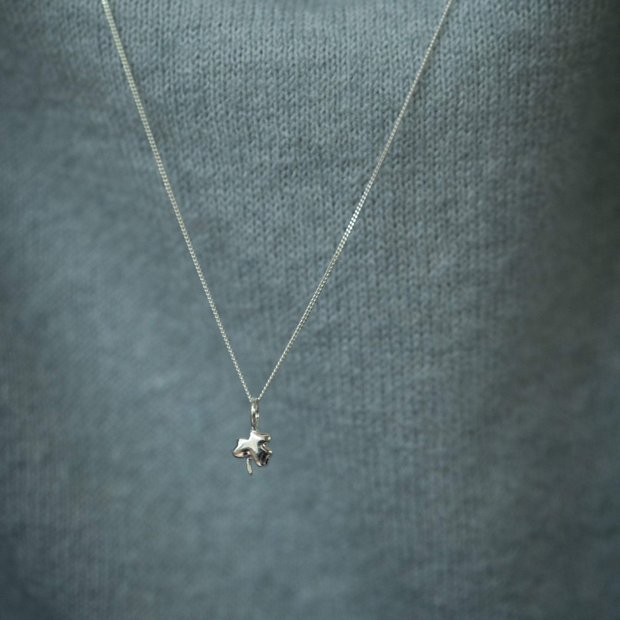 A close-up of a delicate silver chain necklace with a small, detailed shamrock pendant worn by a person wearing a textured slate blue-gray knit sweater.