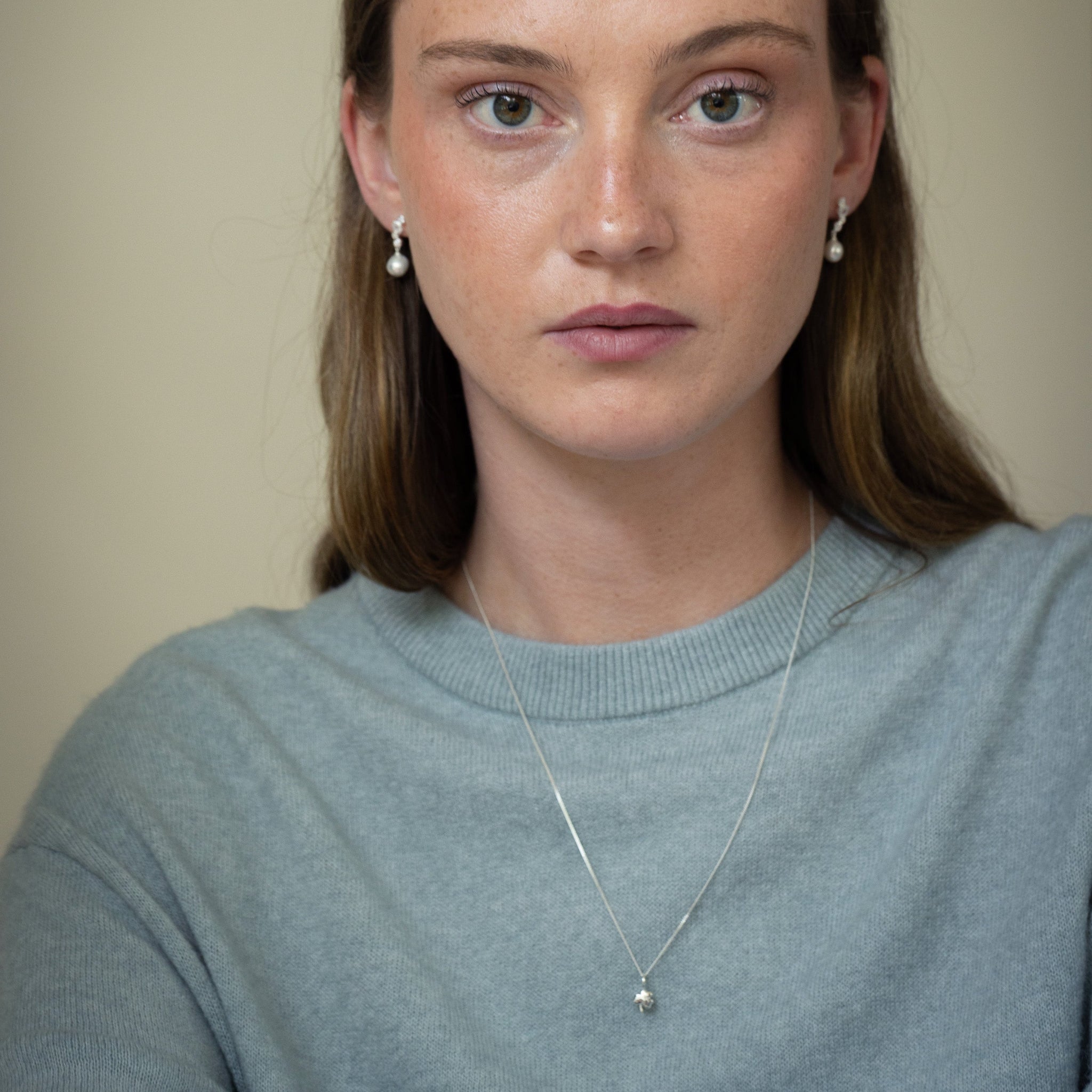 A close-up portrait of a woman with brown hair wearing a light gray knit sweater, a delicate silver necklace, and small pearl-like stud earrings against a plain beige background.
