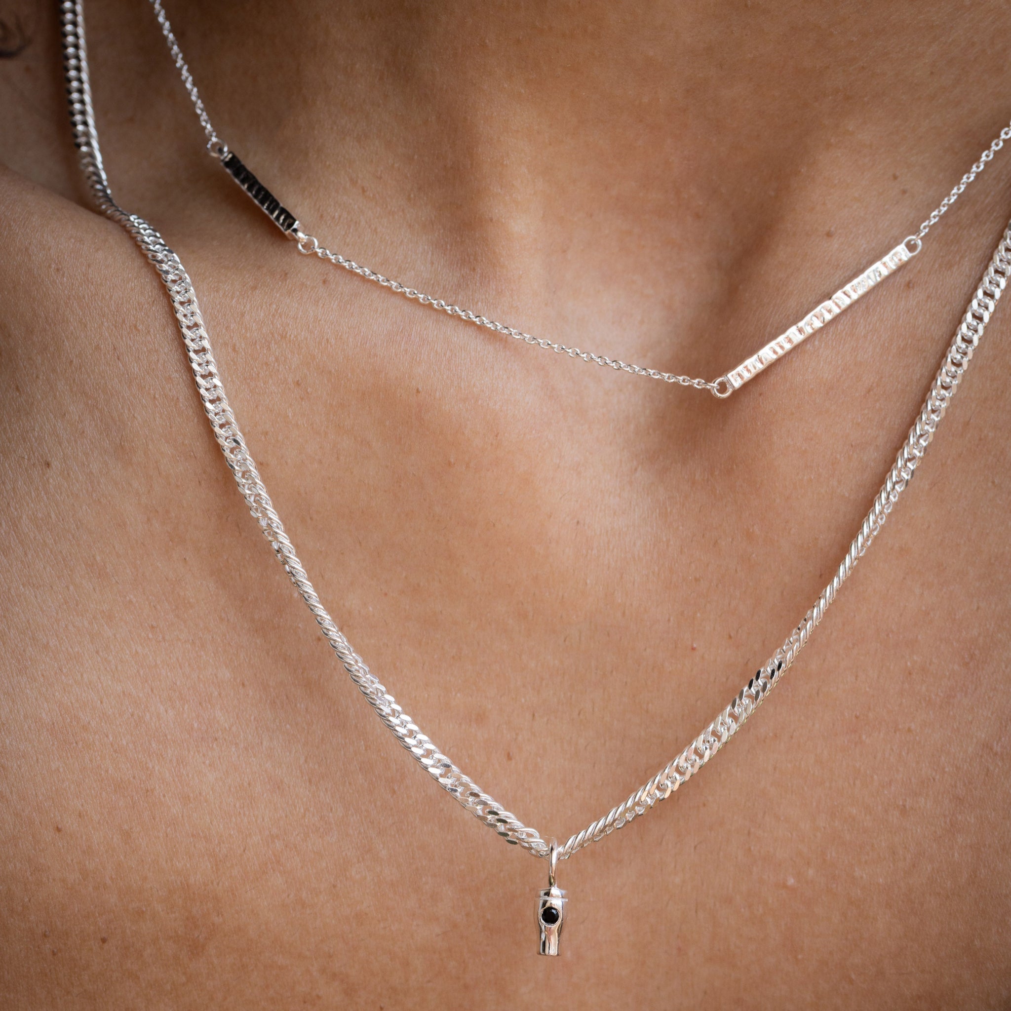 A close-up of a person's neck and upper chest wearing two layered silver necklaces: a curb chain necklace with a small dangling charm and a thinner bar necklace with a small pint shaped pendant.