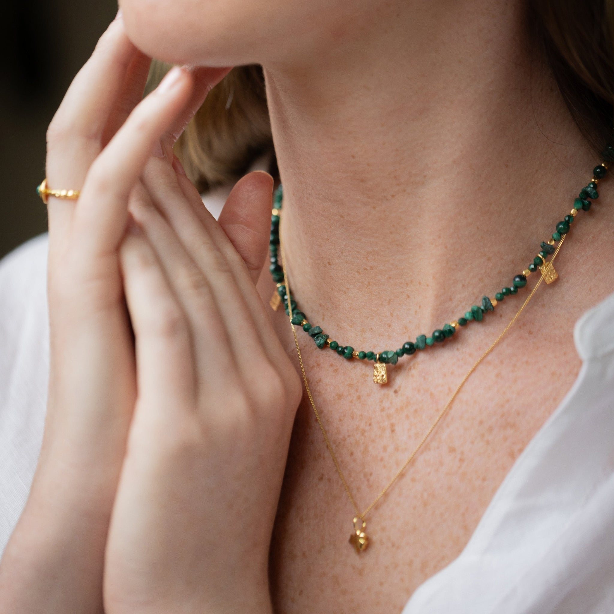 Close-up of a woman wearing a beaded green and gold necklace with matching drop earrings and a delicate gold chain necklace, paired with a white top.