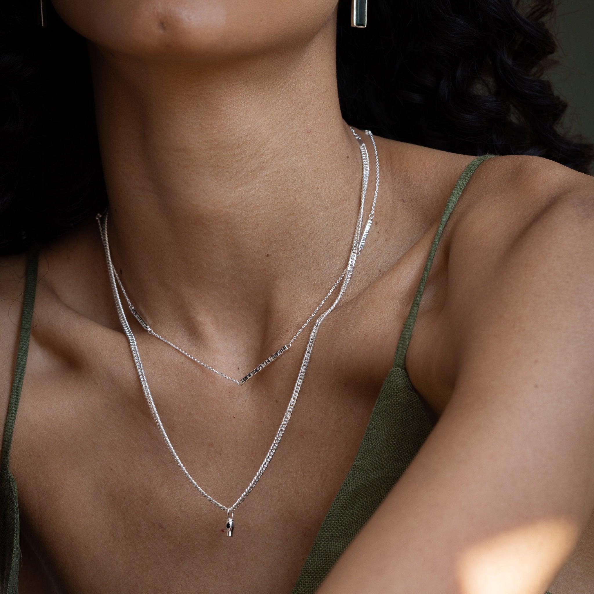 A close-up lifestyle shot of a model wearing layered silver necklaces, including a delicate chain with a small teardrop pendant, layered over a textured chain, paired with a matching silver drop earring.
